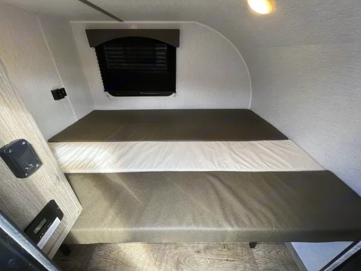 Explore a cozy RV rental interior: a modular bed with dark and white cushions, window, and controls for your trip.. Sunset Park RV SunRay 2023
