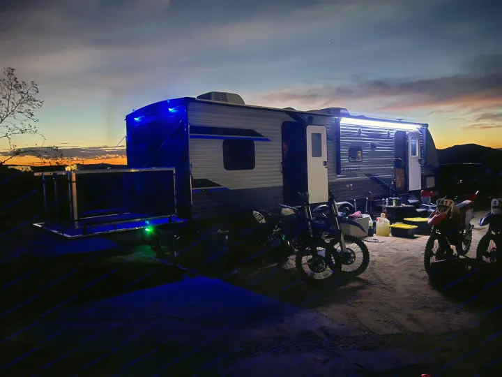 An RV rental glowing with bright lights, set up with dirt bikes for desert camping under a colorful sunset sky.. Coachmen Catalina 2022