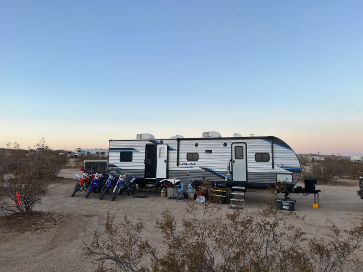 An RV rental toy hauler and several dirt bikes parked in a desert camp, hinting at off-road adventures.. Coachmen Catalina 2022