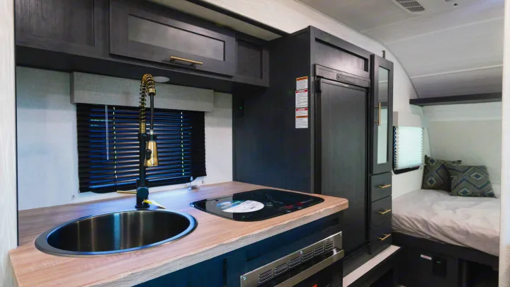 RV rental interior: A compact kitchen with modern dark cabinets, wood counter, sink, and stove, extending to a cozy bed area.. Forest River R-Pod 190 2024
