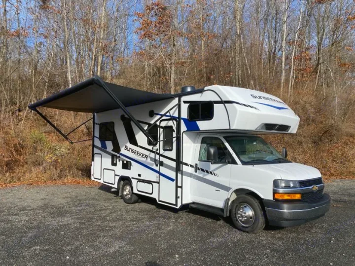 A white Sunseeker RV rental, a Class C model with an extended black awning, is parked on a gravel lot before bare trees.. Forest River Sunseeker 2025