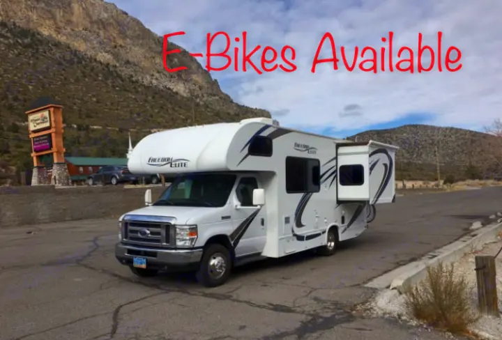 White RV rental with 'E-Bikes Available' text overlay, parked against a scenic mountain backdrop.. Thor Freedom Elite 2018