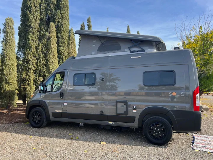Grey Winnebago Solis pop-top camper van, an RV rental, with its roof extended, parked on gravel.. Winnebago Solis BUT59P 2024