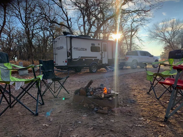 Sunny RV rental camp setup with a Geo Pro travel trailer, pickup truck, chairs, and a warm campfire in the woods.. Forest River Rockwood Geo Pro 2024