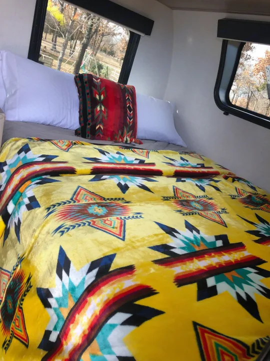 Inside an RV rental: a bed with bright yellow blanket & patterned pillows, offering serene nature views from its window.. Forest River Rockwood Geo Pro 2024