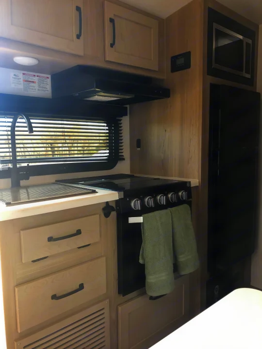 A bright RV rental kitchen showing a black stove, oven, microwave, fridge, sink, and light wood cabinetry. Towels hang on the oven handle.. Forest River Rockwood Geo Pro 2024