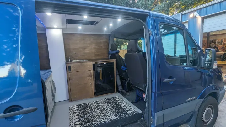 This blue RV rental Sprinter van interior features a compact kitchen with sink, fridge, and a bed, ideal for travel.. Dodge Sprinter 2007