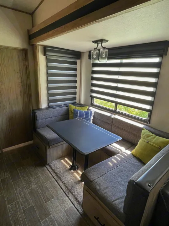 RV rental dinette with grey booth seating, dark table, and striped blinds. Sunlit interior of a camper.. Forest River Cherokee Arctic Wolf 2021