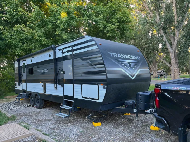 A Transcend XPLOR travel trailer (RV rental) hitched to a 4x4 truck is parked at a gravel campsite among trees.. Grand Design Transcend Xplor 265BH 2022