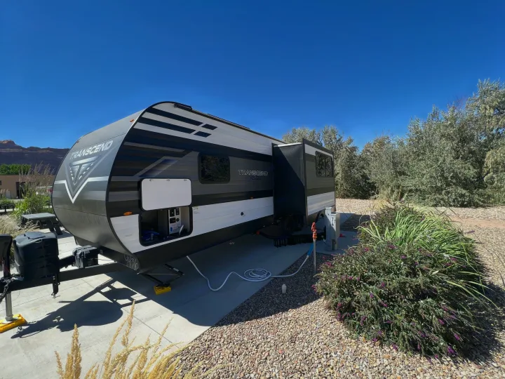 An RV rental, a Transcend travel trailer, set up at a sunny campsite with hookups, surrounded by desert landscaping.. Grand Design Transcend Xplor 265BH 2022