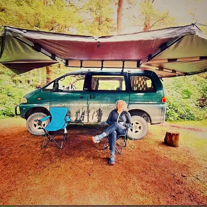 A smiling woman relaxes in a camp chair beside her green, awning-covered RV rental in a forest clearing, ready for adventure.. MITSUBISHI Delica 1997