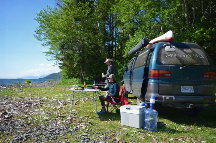 Two people enjoy outdoor cooking next to their dark-colored RV rental van by a scenic waterfront with trees under a clear sky.. MITSUBISHI Delica 1997