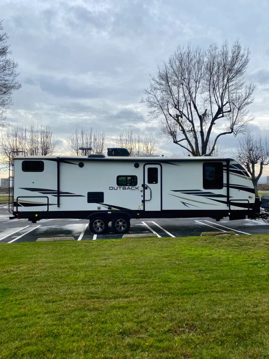 White Outback travel trailer, great for RV rental, parked on asphalt. Overcast sky & bare trees in background.. Keystone RV Outback Ultra-Lite 2022