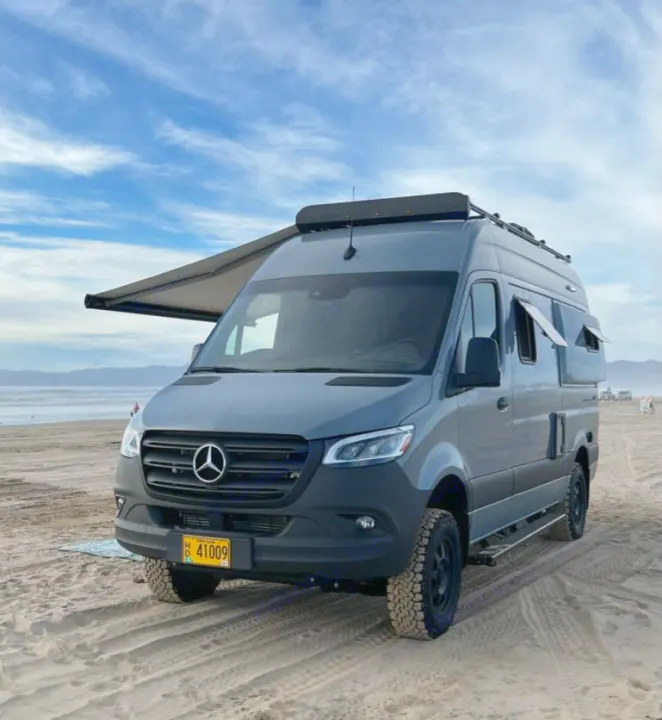 A grey Mercedes Sprinter RV rental van is parked on a sandy beach. Its awning is extended, with the ocean visible in the background.. Winnebago Revel 2022