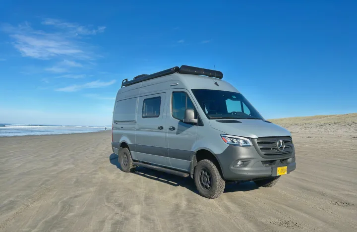 Grey Mercedes Sprinter camper van (RV rental) on a sandy beach by the ocean. A person walks in the distance. Perfect for travel.. Winnebago Revel 2022