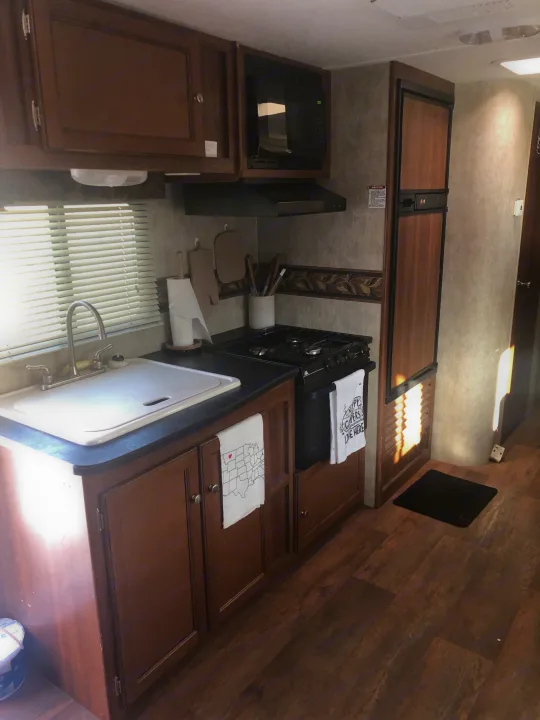 RV rental kitchen: compact space with wood cabinets, sink, gas stove, microwave, and fridge. Sunlight brightens the interior.. Keystone RV Bullet Ultra Lite 2015