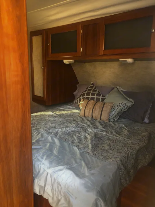 Inside a cozy RV rental bedroom featuring a bed with patterned bedding, decorative pillows, and overhead wooden storage cabinets.. Keystone RV Bullet Ultra Lite 2015