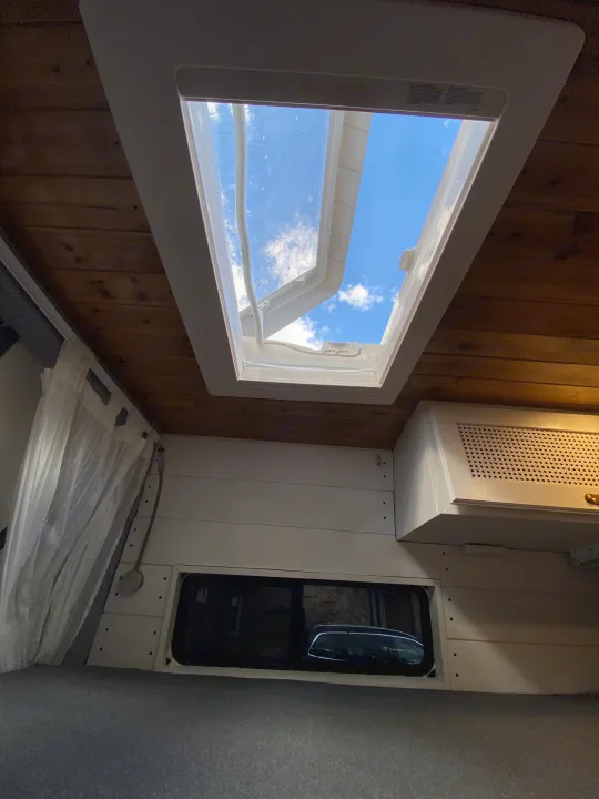 Cozy RV rental interior: open skylight reveals blue sky, wooden ceiling, white paneled walls, and a comfortable bed.. Mercedes Sprinter Premium 2020