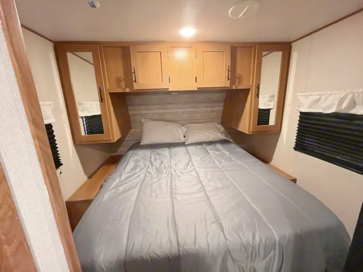 Cozy RV rental bedroom with a comfortable bed, wood cabinets, and window blinds. Ideal for travel.. Forest River Prime Time Avenger 27 DBS 2023