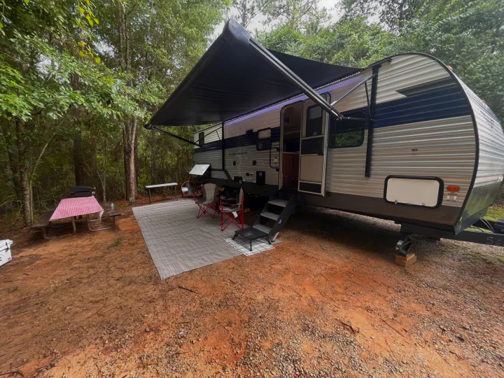 Spacious RV rental set up at a wooded campsite with awning, outdoor rug, and picnic area. Perfect for a nature getaway!. Forest River Prime Time Avenger 27 DBS 2023
