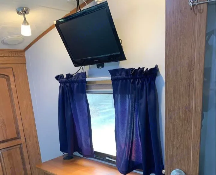 RV rental interior view: A small TV is mounted above a window with dark blue curtains, flanked by wood cabinetry.. Forest River Palomino Sabre 2015