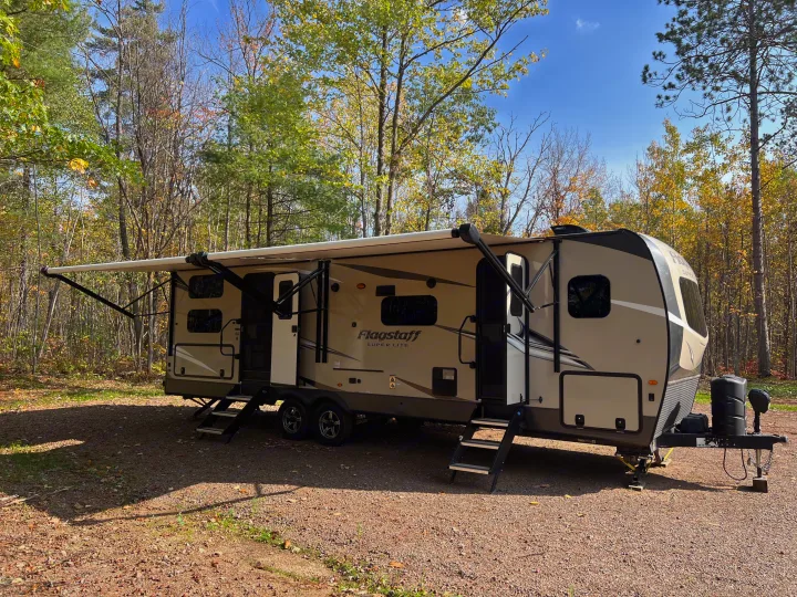 A beige Flagstaff travel trailer RV rental with an awning extended, parked on gravel in a colorful autumn forest campsite.. Forest River Flagstaff Super Lite 2021