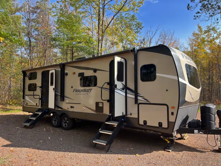 RV rental: A tan travel trailer camper with multiple pop-outs extended, parked on gravel in a sunny wooded area.. Forest River Flagstaff Super Lite 2021