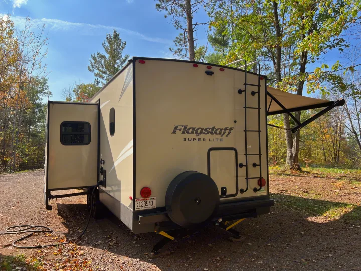 Rear view of a tan Flagstaff travel trailer, likely an RV rental, set up at a leafy fall campsite with its slide-out and awning open.. Forest River Flagstaff Super Lite 2021