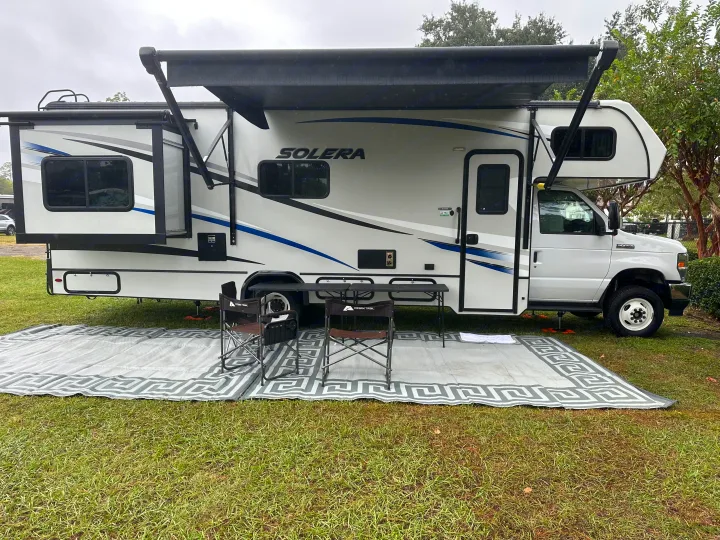 Spacious rv rental parked at a serene campsite, featuring an extended awning, outdoor mat, chairs, and table for comfortable outdoor living.. Forest River Solera 2021