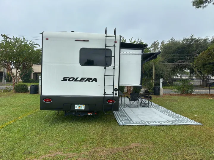 Solera RV rental parked for camping, featuring a large slide-out, ladder, and outdoor living area with mat and chairs.. Forest River Solera 2021