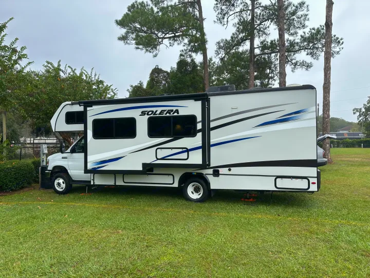 White Solera RV rental parked on green grass at a campsite, with its slide-out extended, surrounded by trees.. Forest River Solera 2021