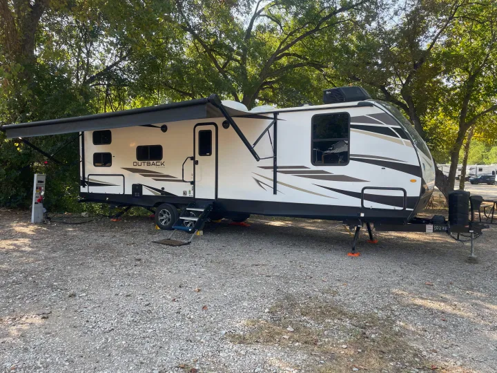 RV rental: A white travel trailer is parked on a gravel site with its awning extended under lush leafy trees.. Keystone Outback 2022
