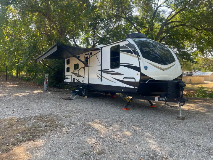 A white Outback travel trailer, an RV rental, set up with an awning extended on a gravel campsite amidst lush trees.. Keystone Outback 2022