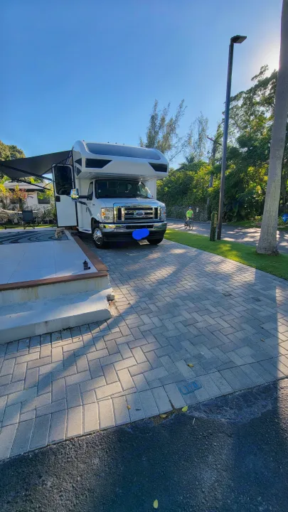 A white RV rental is parked on a spacious, sunny paved site. A person on a bicycle rides by in the background.. Jayco Greyhawk 2023