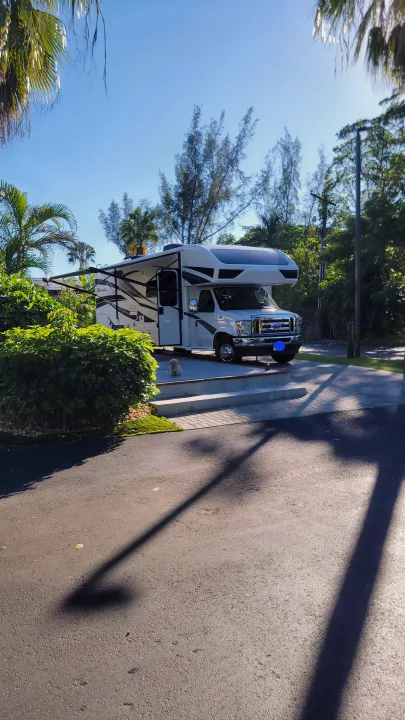 White RV rental with awning extended, parked on a sunny paved lot amidst green trees. A small white dog sits nearby.. Jayco Greyhawk 2023