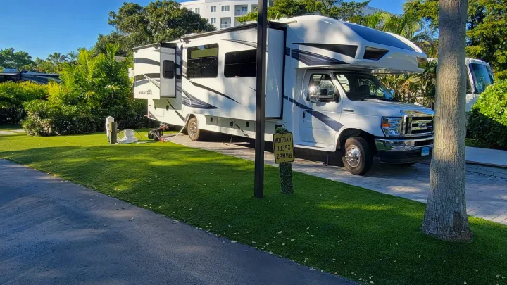 A white RV rental is parked on a paved site at a sunny resort, featuring green turf, palm trees, and an upside-down sign.. Jayco Greyhawk 2023