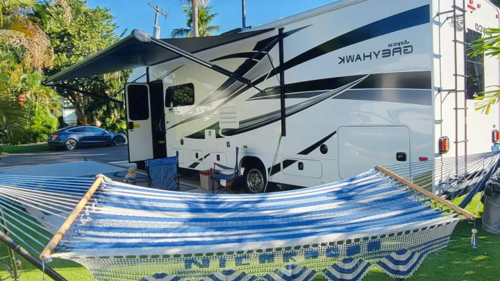 White Jayco Grayhawk rv rental setup with a striped hammock, chairs, and a dog. Palm trees and a Tesla are in the background.. Jayco Greyhawk 2023
