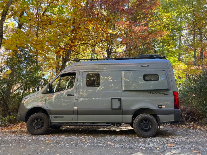 Gray Winnebago Revel 4x4 camper van on a gravel path, surrounded by vibrant autumn trees. Perfect RV rental.. Winnebago Revel 2023