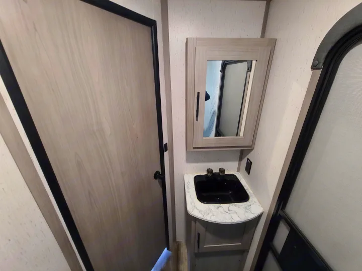Compact RV rental bathroom with light wood door, black sink on a marble-look counter, and a mirrored cabinet above.. Dutchmen Coleman 2022