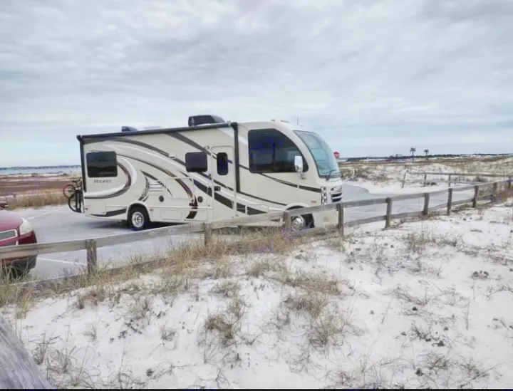An RV rental motorhome is parked on a paved lot beside white sand dunes and a coastal body of water.. Thor Vegas 2016