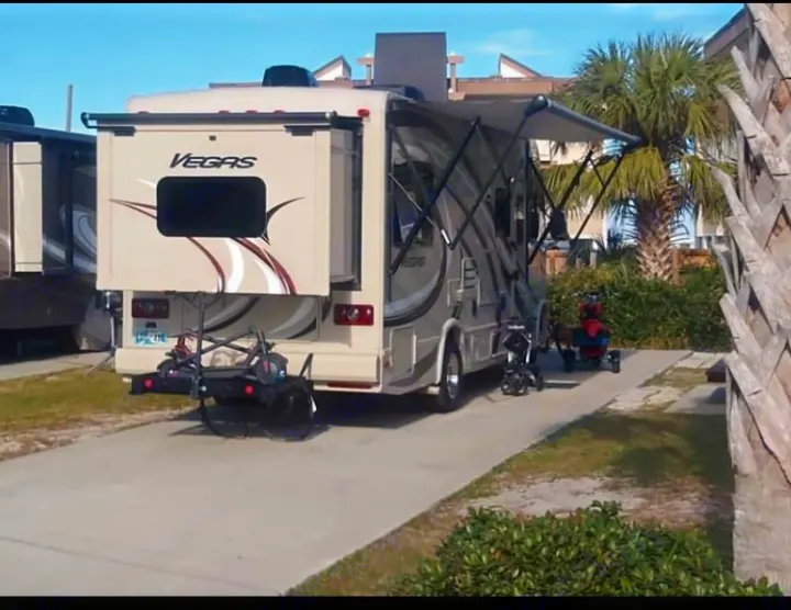 Beige Vegas RV rental parked with slide-out and awning extended. Scooter and mobility aid carriers are attached at a sunny campground.. Thor Vegas 2016