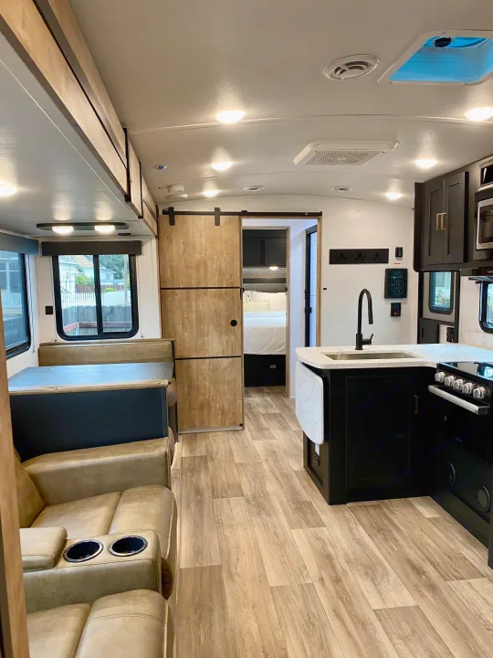 Spacious RV rental interior featuring a modern kitchen, dining booth, comfy recliners, and a bedroom beyond a barn door.. Keystone RV Outback Ultra-Lite 2022