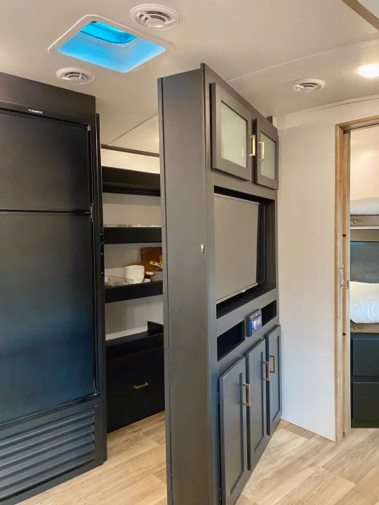 RV rental interior: black fridge, pantry, and a modern grey entertainment unit dividing living and bunk areas.. Keystone RV Outback Ultra-Lite 2022