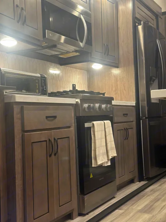 Step inside this sleek RV rental kitchen. Features a microwave, range, refrigerator, and light wood cabinets.. Cedar Creek Cedar Creek Fifth Wheel 2022
