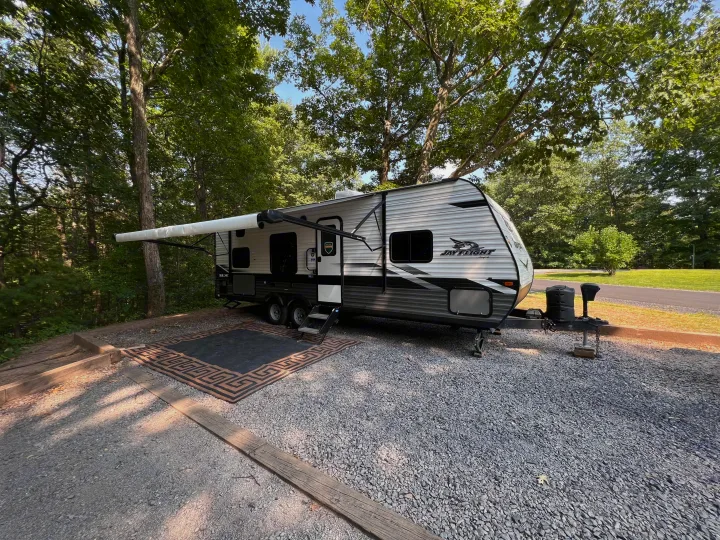 An RV rental with its awning extended and a rug laid out, parked at a spacious gravel campsite amidst lush trees.. Jayco Jay Flight SLX 2022