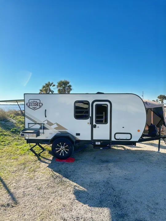 This RV rental is set up by the water, featuring an outdoor griddle for camping fun under a clear blue sky.. Dutchmen Other 2023