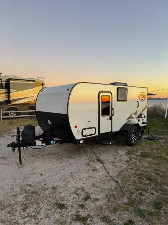 This compact RV rental, a white Rubicon travel trailer, glows at sunset by the water, perfect for outdoor camping adventures.. Dutchmen Other 2023