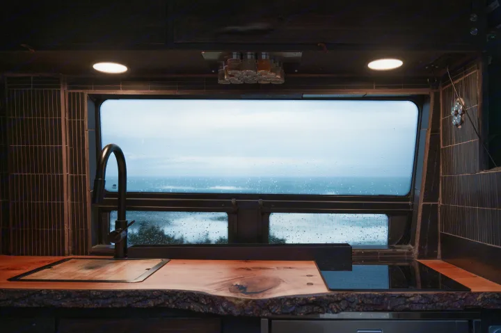 RV rental: Rustic kitchen with wood countertop, sink, and stovetop. Ocean view through rain-splattered window; lights on.. Mercedes-Benz Sprinter 2021