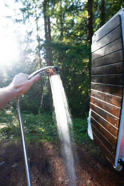 Hand holding chrome shower head spraying water in a sun-dappled forest, part of an RV rental outdoor setup.. Mercedes-Benz Sprinter 2021