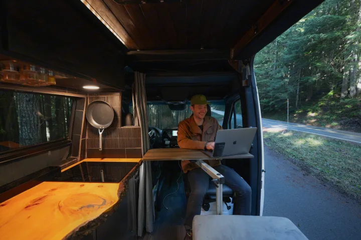 Man smiles, working from his RV rental mobile office, parked roadside by a forest. Ideal for digital nomads.. Mercedes-Benz Sprinter 2021
